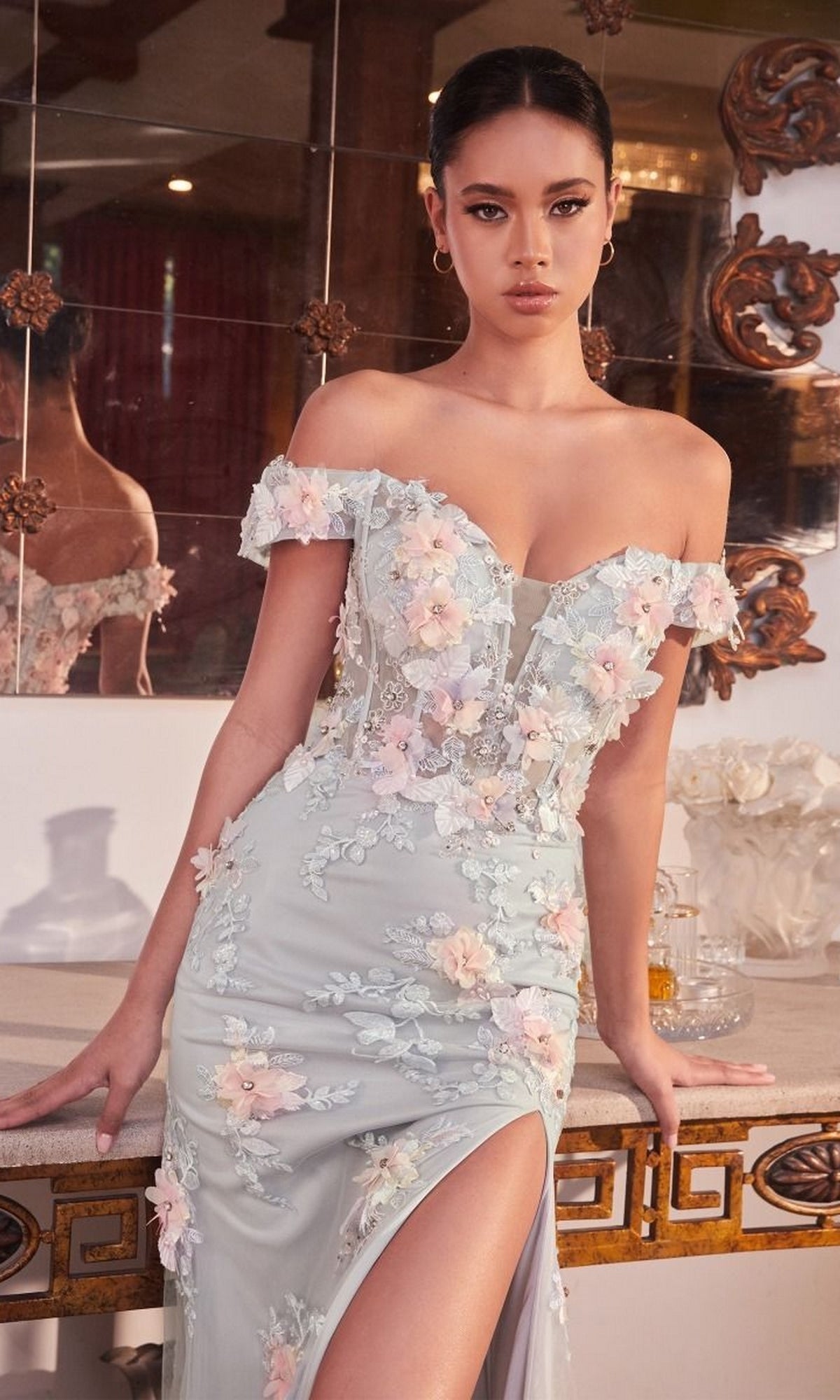 Woman wears the Floral-Embellished Long Mist Gray Prom Dress CD005, featuring an off-the-shoulder design, floral appliqués, and a thigh-high slit, standing indoors by a decorative mirror.